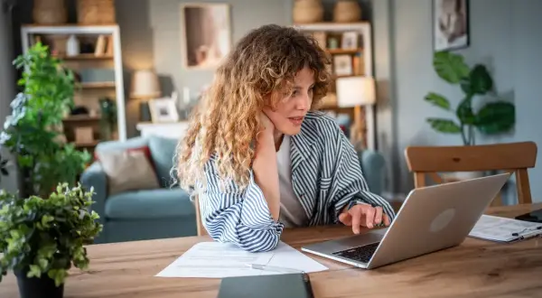 Donna con capelli ricci lavora al computer, documenti sul tavolo. Forse sta cercando informazioni su un mutuo con garante genitore per realizzare un sogno.
