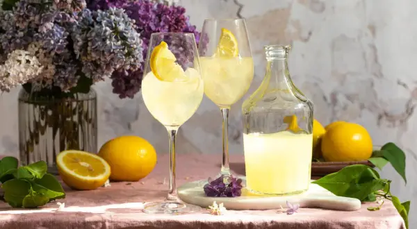 Zwei Gläser Limoncello Spritz mit Zitrone, eine Flasche Limoncello, Zitronen und Flieder auf rosafarbenem Tischtuch.