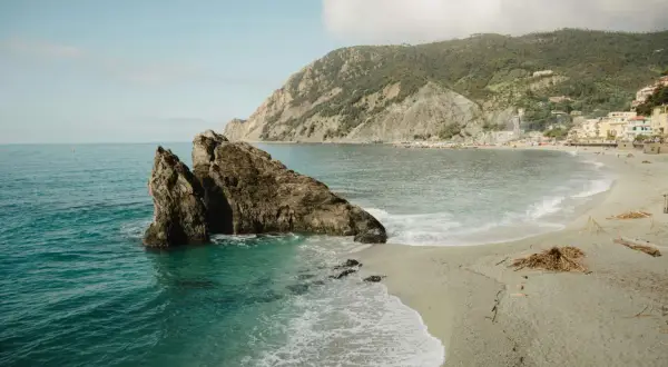 Un rocher émerge des eaux turquoise d'une crique. Une plage de galets s'étend vers un village coloré niché sur une colline verdoyante. Idéal pour savoir où partir en vacances cet été.