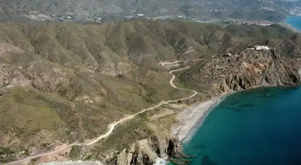 Vista aérea de la playa el lance, con su arena dorada, aguas turquesas y acantilados rocosos. Un camino serpentea por las colinas áridas.