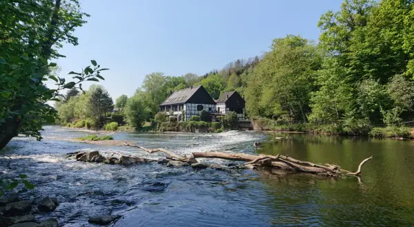Fachwerkhäuser am Flussufer, wo die **Wipperkotten wandern**. Ein Baumstamm liegt im rauschenden Wasser.