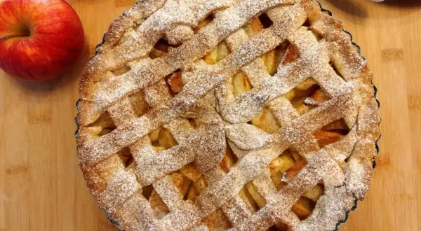 Pyszna tarta z jabłkami, posypana cukrem pudrem, obok leżą świeże jabłka.