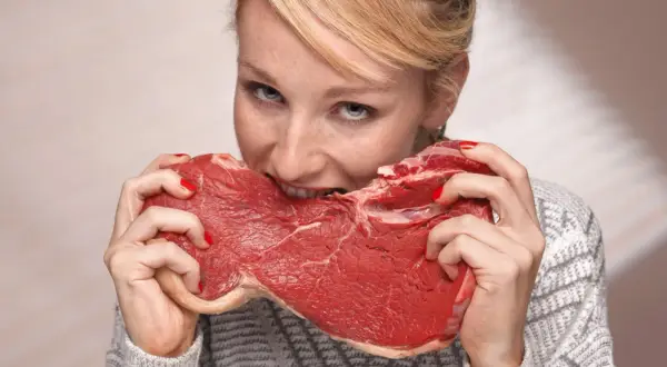 Kobieta z rudymi włosami i piegami wgryza się w surowy kawałek mięsa, jakby to była jej dieta lwa.