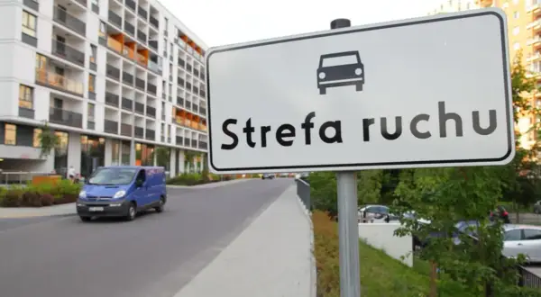 Znak "Strefa ruchu" przed nowoczesnym budynkiem mieszkalnym. Niebieski bus jedzie po drodze.