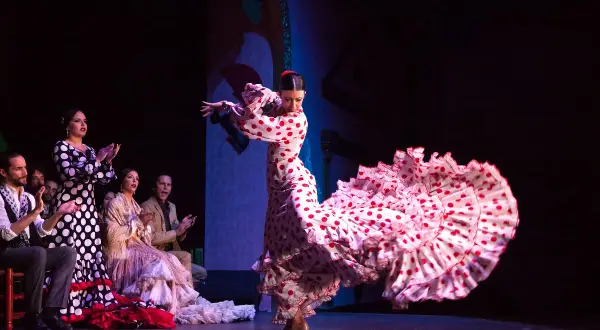 Bailarina de flamenco con un vestido de lunares rojos y blancos, su falda ondea con gracia. El público aplaude la seguidilla flamenca.