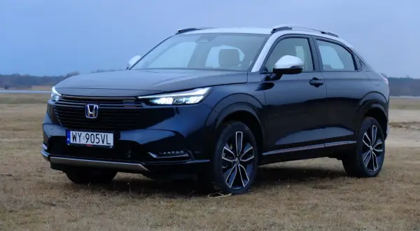 Nowa Honda HR-V w ciemnym kolorze, zaparkowana na trawiastym polu, z widokiem na jezioro i las w tle.