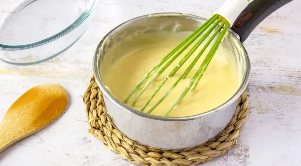 Un batidor verde mezcla una crema amarilla en una olla, mostrando cómo espesar un puré. Cerca hay una cuchara de madera y un recipiente de vidrio.