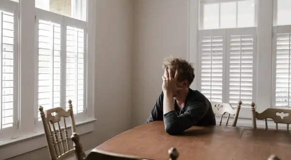 Hombre con la mano en la cara, sentado a una mesa de madera, frente a una ventana de tolerancia con persianas blancas.