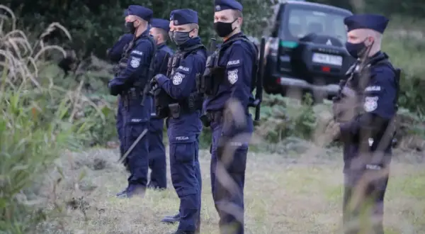 Policjanci w pełnym umundurowaniu, w tym w maskach, stoją w gotowości. W tle widać radiowóz. Sytuacja wygląda na stan wyjątkowy.