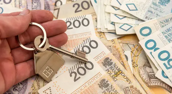 Dłoń trzyma klucz z breloczkiem w kształcie domu na tle polskich banknotów. Symbolizuje to możliwość zakupu nieruchomości lub dziedziczenia, co wiąże się z kwestią zachowku.
