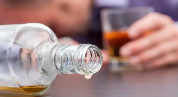 Ostatnia kropla alkoholu metylowego kapie z butelki. W tle niewyraźna postać trzyma szklankę z bursztynowym płynem.