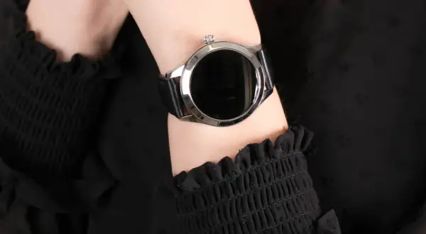 Damski smartwatch z czarnym paskiem na nadgarstku, na tle czarnej bluzki.