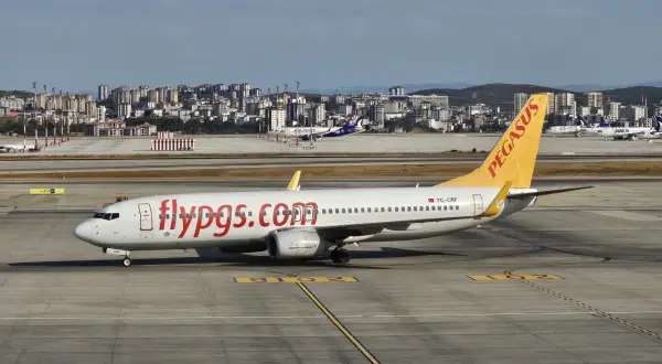 Samolot linii lotniczych Pegasus z napisem "flypgs.com" na kadłubie na płycie lotniska, w tle miasto.