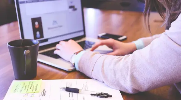 Kobieta pracuje przy laptopie. Na biurku kubek, notatki i długopis. Oświetlenie zapewnia odpowiednie ile luxów na stanowisku pracy.