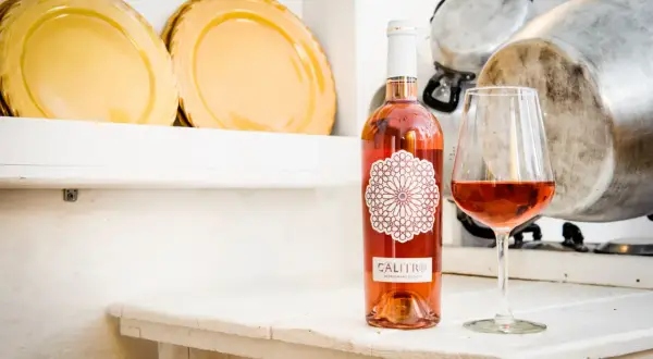 Un bicchiere e una bottiglia di negroamaro rosato Calitro su un tavolo bianco, con piatti gialli e pentole sullo sfondo.