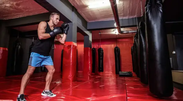 A boxer trains in a gym with many boxing bags. The red floor reflects the lights, and the boxing bag numbers are not visible.