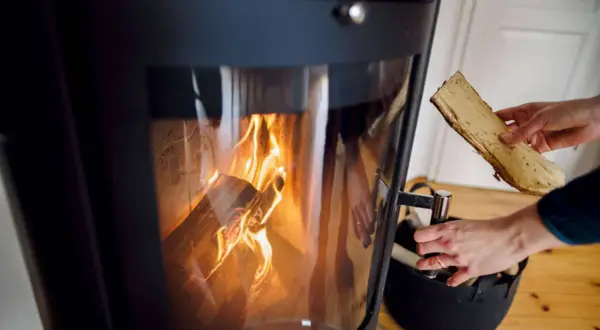 Eine Hand legt Holzscheite in einen brennenden Ofen. Die Flammen lodern im Inneren des Ofens.