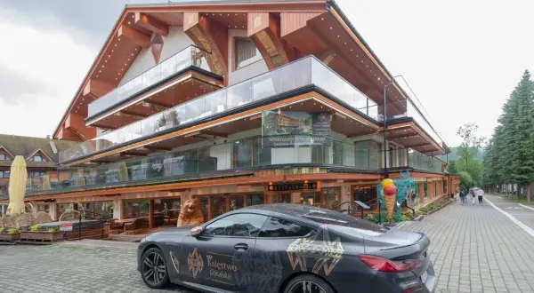 Luksusowy hotel Górski Diament z nowoczesną architekturą i przeszklonymi balkonami. Przed budynkiem stoi elegancki samochód z motywem "Księstwo Góralskie".