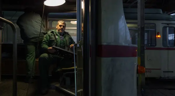 Mężczyzna w zielonym kombinezonie siedzi w kabinie tramwaju, trzymając panel sterowania. To fotografia miejska z wnętrza zajezdni.