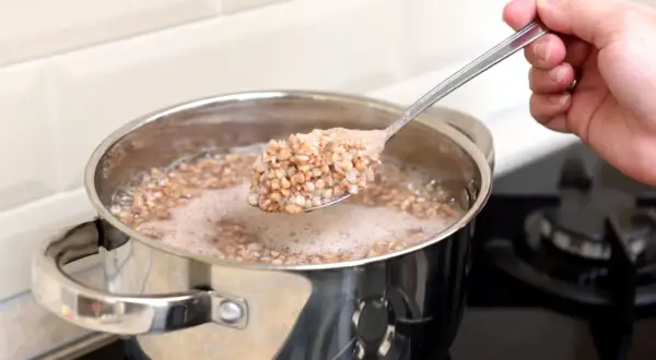 Eine Hand hebt einen Löffel mit dampfendem Buchweizen aus einem Topf, in dem Buchweizen gekocht wird.