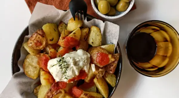 Patatas bravas con alioli, servidas con aceitunas y una bebida oscura.