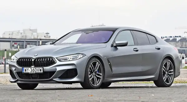 Szary BMW M8 Gran Coupe z czarnym dachem i felgami, zaparkowany na ulicy.