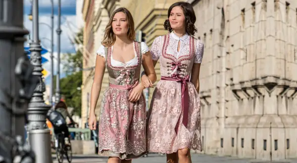 Zwei Frauen in Dirndln ohne Schnürung spazieren. Die Kleider sind in Rosa- und Beigetönen gehalten und mit zarten Stickereien verziert.