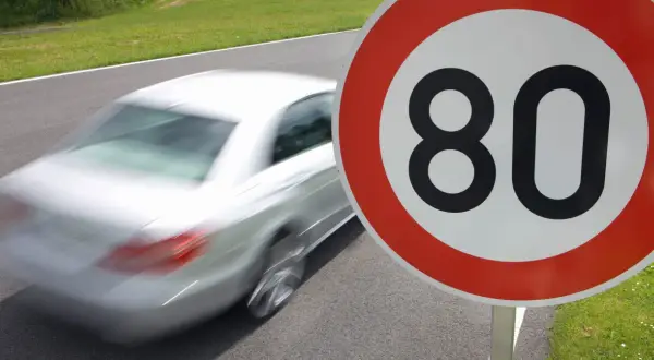Znak ograniczenia prędkości do 80 km/h. Biały samochód pędzi po drodze, przypominając o przepisach ruchu drogowego i ograniczeniach prędkości w Polsce.