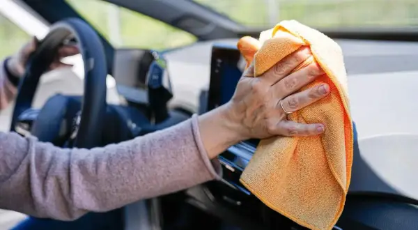 Mano limpiando el salpicadero del coche con un paño naranja. Descubre trucos caseros para limpiar el salpicadero del coche y mantenerlo impecable.