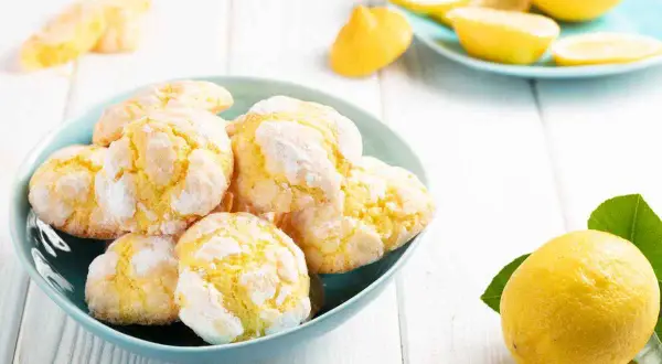 Biscotti al limone, morbidi e profumati, in una ciotola azzurra, con limoni freschi e spicchi su un piattino sullo sfondo.