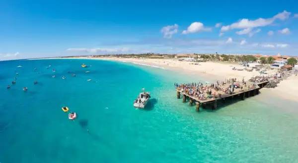 Tłum ludzi na molo, łodzie na turkusowym morzu, plaża i budynki. Zastanawiasz się, ile się leci na Wyspy Zielonego Przylądka?