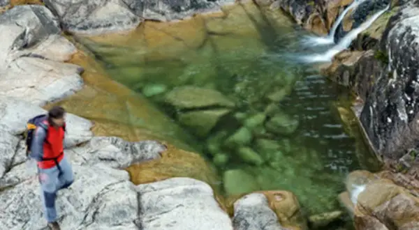 Senderista se detiene junto a un **río caldo** de aguas cristalinas y rocas musgosas.