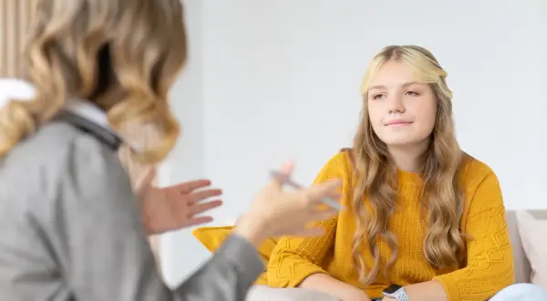 Nastolatka w żółtym swetrze podczas terapii. Rozmowa z terapeutką, która gestykuluje.