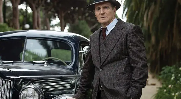 Philip Marlowe, detektyw z kryminałów Raymonda Chandlera, w eleganckim garniturze i kapeluszu, opiera się o luksusowy samochód z lat 40.