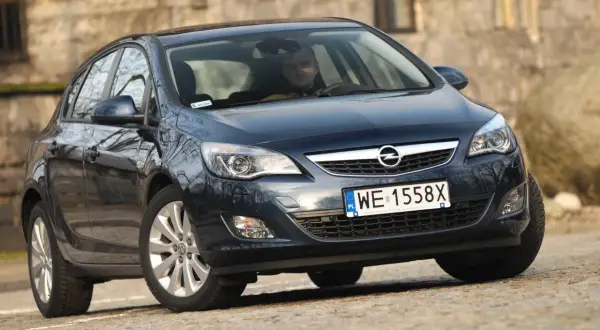 Ciemny Opel Astra J z włączonymi światłami do jazdy dziennej, zaparkowany na kamienistej drodze.