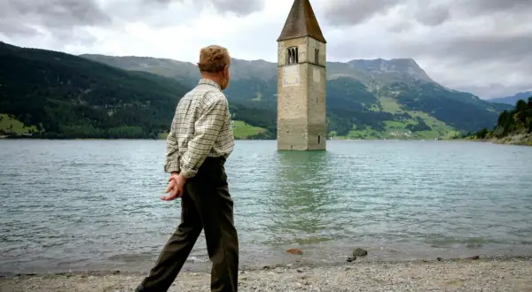 Ein Mann spaziert am Ufer eines Sees, auf dem ein Kirchturm aus dem Wasser ragt. Die Kirche im Stausee ist ein eindrucksvolles Zeugnis der Vergangenheit.