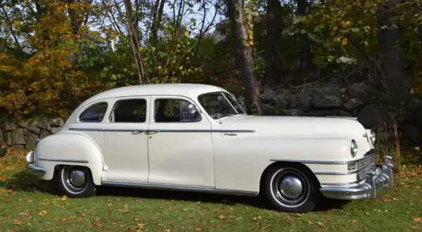 En vit Chrysler New Yorker från 1940-talet står parkerad på en gräsmatta med höstlöv.