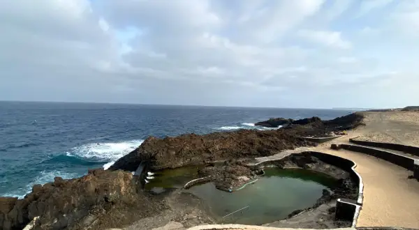 Piscinas naturales formadas por lava volcánica en la costa de Lanzarote, con el mar azul de fondo. No hay cocoteros, pero sí un paisaje salvaje.