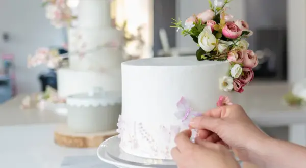 Una mano aggiunge una farfalla a una torta bianca decorata con fiori e paillettes. Sullo sfondo, un'altra torta a più piani con decorazioni floreali rosa.