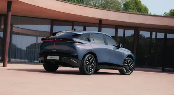 Lancia Gamma 2026, un'elegante berlina blu metallizzato con fari posteriori a LED, parcheggiata davanti a un edificio moderno.