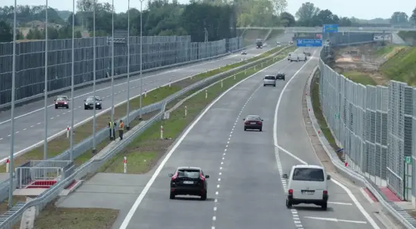 Samochody poruszają się lewym pasem w terenie zabudowanym, mijając ekrany akustyczne.