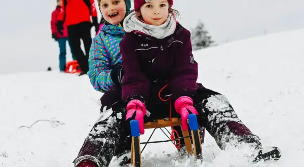 Zwei Kinder rodeln lachend einen verschneiten Hang im Bergischen Land hinunter.