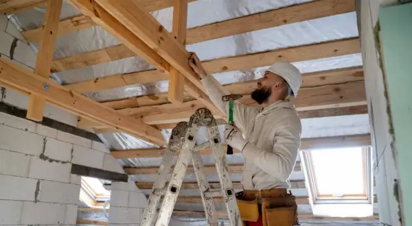 Obrero con casco y martillo realiza refuerzo estructural de vigas de madera en una construcción.