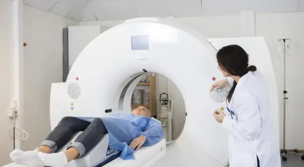 Pacjentka leży na stole tomografu, przygotowana do badania. Lekarka w białym fartuchu obsługuje aparat, który wykonuje angiografię.