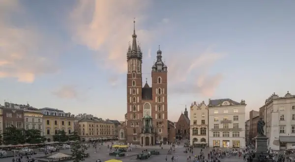 Krakowski Rynek Główny z majestatyczną Bazyliką Mariacką w tle. Ludzie spacerują, a nad nimi pastelowe chmury.