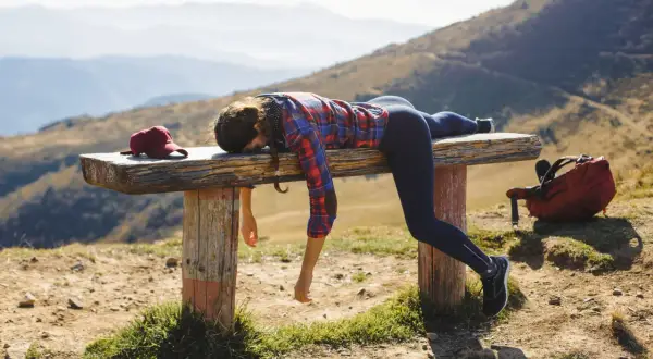 Donna esausta si riposa su una panca in montagna, con zaino e cappellino. Ottimi trekking consigli per non finire così!