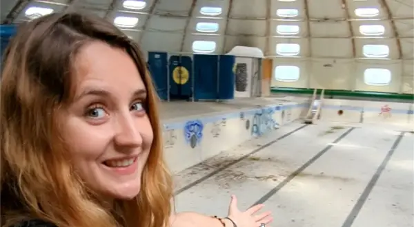 Une jeune femme souriante explore une piscine tournesol abandonnée, son dôme blanc orné de fenêtres rondes.