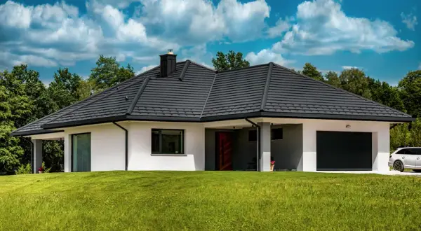 Nowoczesny dom z ciemnym dachem, którego grubość dachówki ceramicznej zapewnia trwałość. Otoczony zielenią, z podjazdem i białymi ścianami.