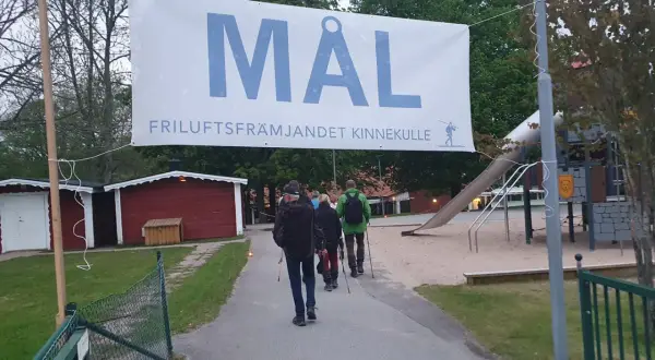 A banner reading "MÅL FRILUFTSFRÄMJANDET KINNEKULLE" hangs over a path where people walk towards a playground.