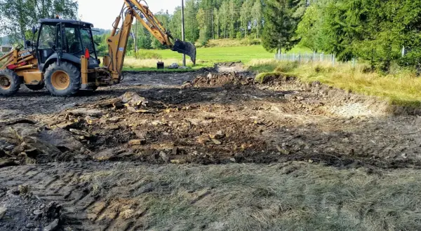 Koparka pracuje, tworząc wykopy pod fundamenty. Teren jest błotnisty, z gruzem i kamieniami.